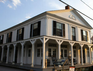 A large white building with many windows and a wrap-around porch and rocking chairs.