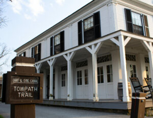 A white building with many windows and a wrap around porch. In front of it is a sign that reads, "Ohio and Erie Canal Towpath Trail."