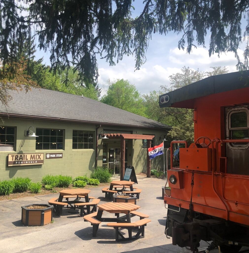A photo of a brick store painted green with a sign that reads Trail Mix Peninsula. Picnic tables and a painted orange train car are in the foreground. 
