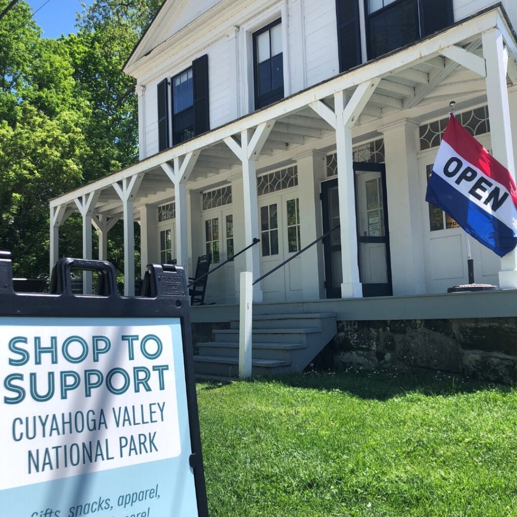 A photo of a large white home with a wrap-around porch and black shutters that now serves as the location for Boston Store. A flag that reads open is out front along with a sign that reads: Shop to support Cuyahoga Valley National Park. 