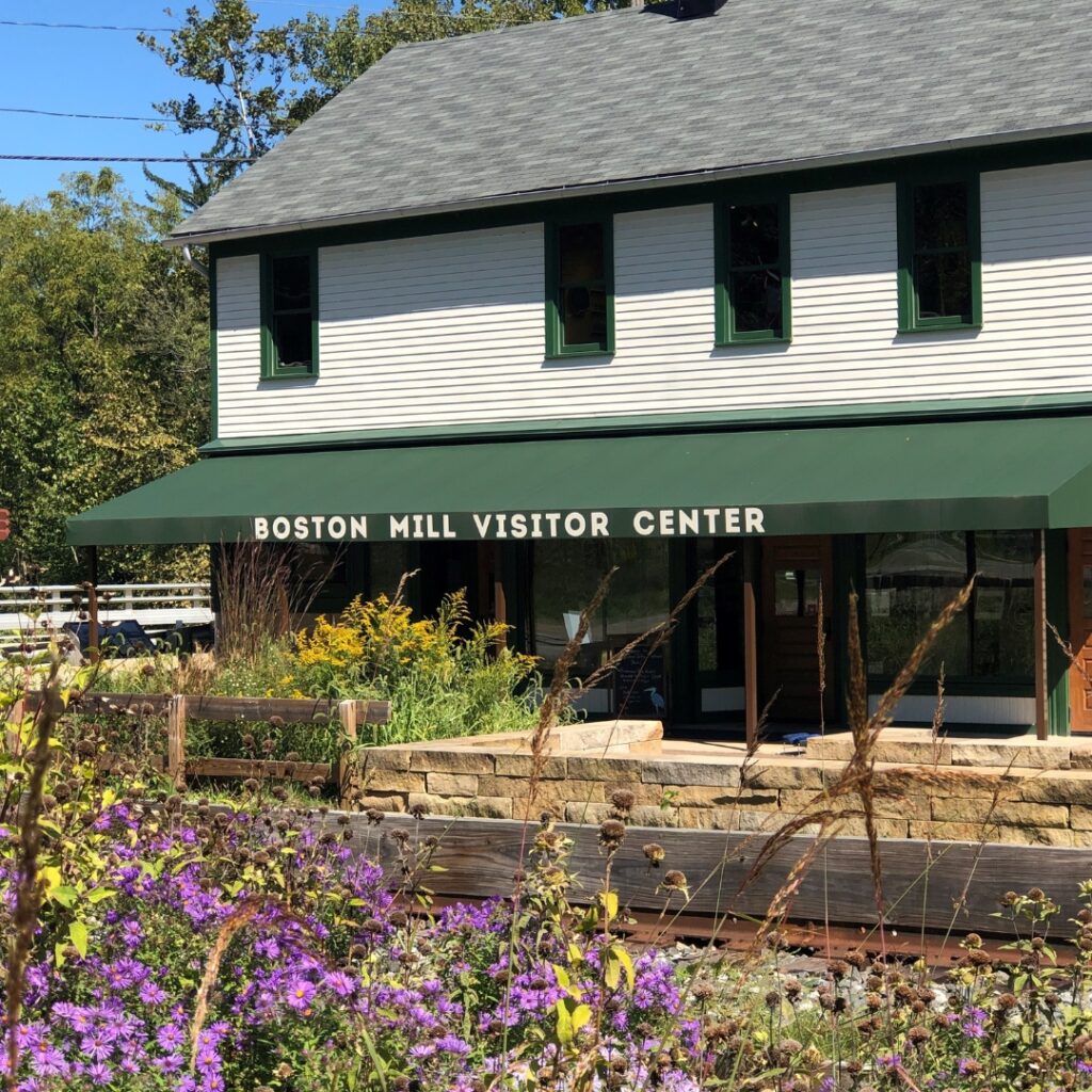 A photo of a two-story sided building with green shutters and a green awning that reads Boston Mill Visitor Center. Plants and flowers are in the foreground. 