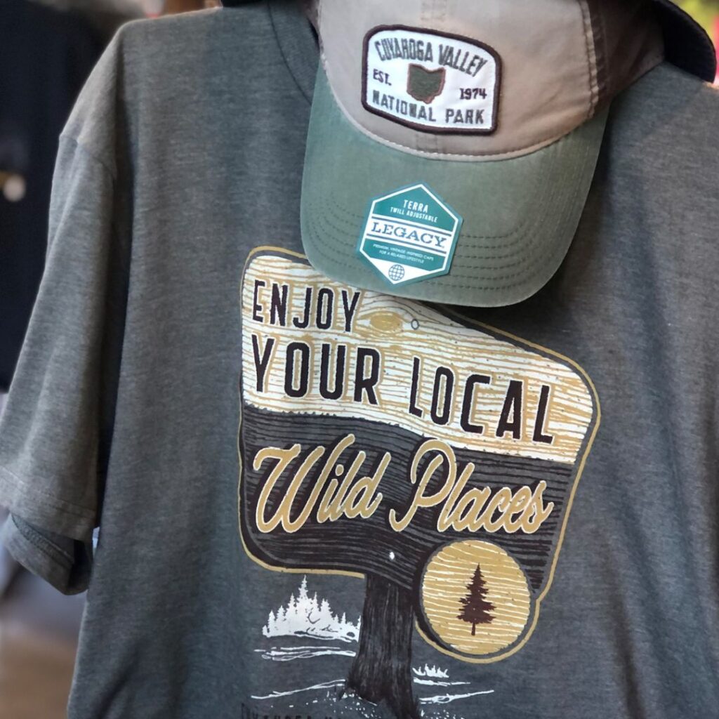 A photo of a gray t-shirt with a graphic illustration of a wooden signpost that reads: Enjoy your local wild places, Cuyahoga Valley National Park. A Cuyahoga Valley National Park baseball hat is also pictured. 