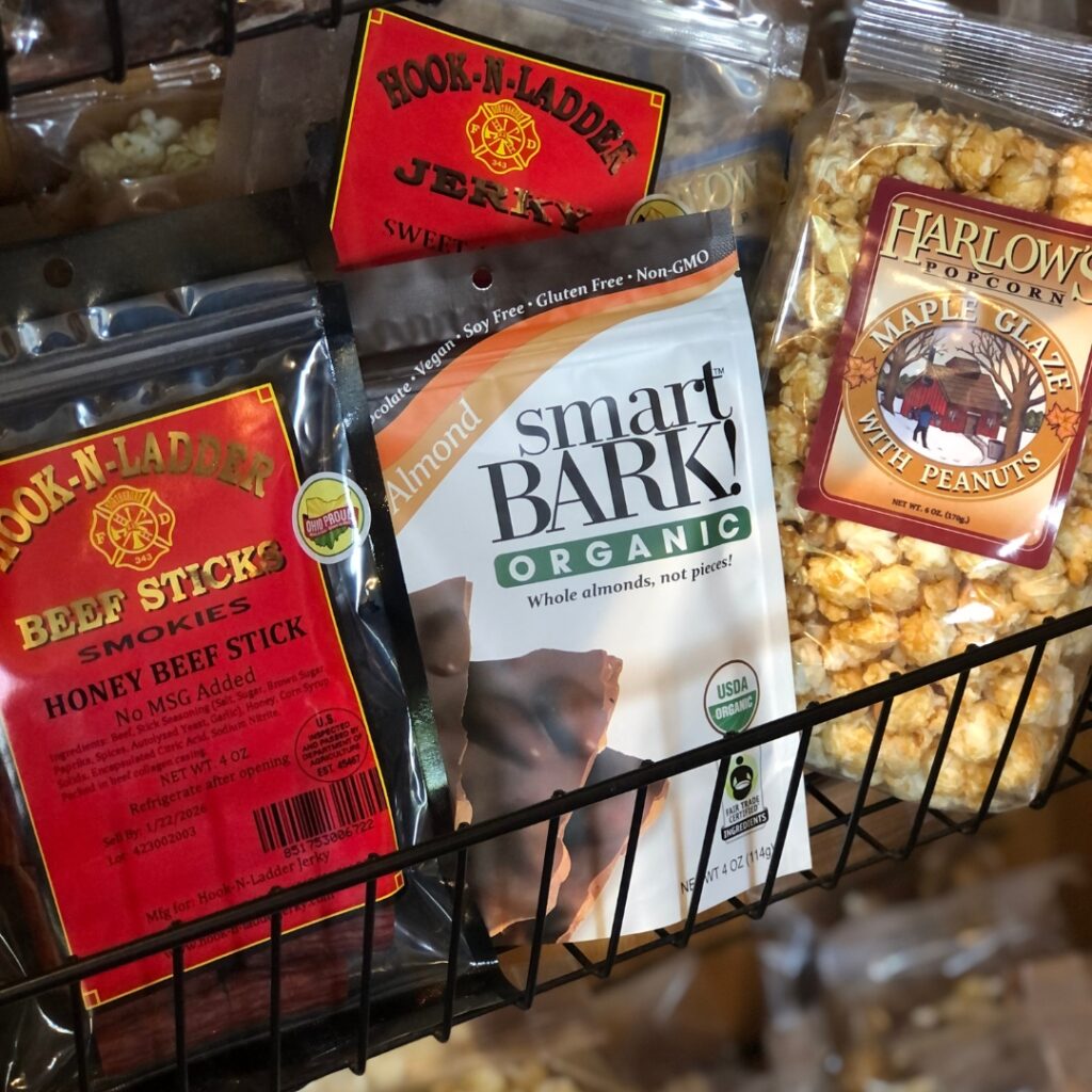 A photo of several snacks in a wire shelf bin. Snacks include, jerky, beef sticks, chocolate bark, and flavored popcorn. 