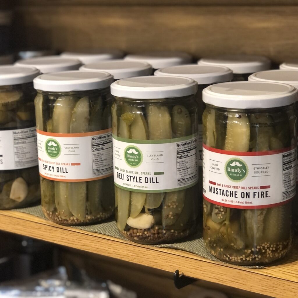A photo of several jars of pickles with flavor names such as spicy dill, deli style dill, and mustache on fire. 