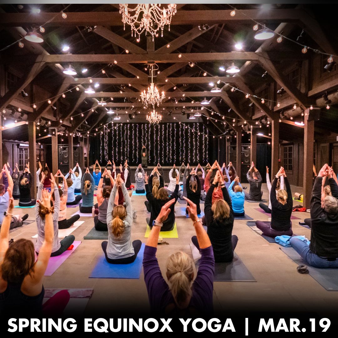 A photo of a large group of people doing yoga poses indoors. The words Spring Equinox Yog, March 19 are at the bottom of the photo.