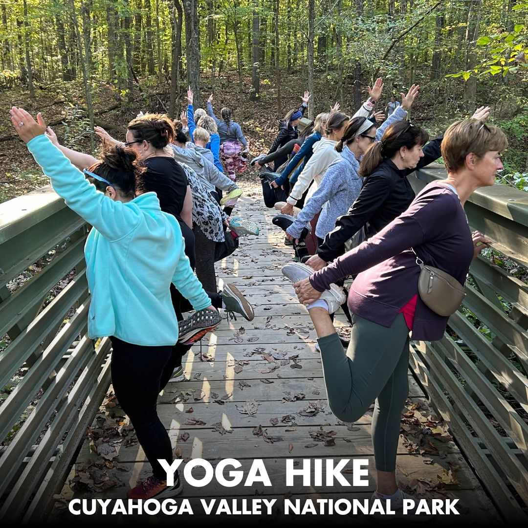 A group of people on a foot bridge connecting to a forest trail hold yoga poses. Text reads: Yoga Hike, Cuyahoga Valley National Park.